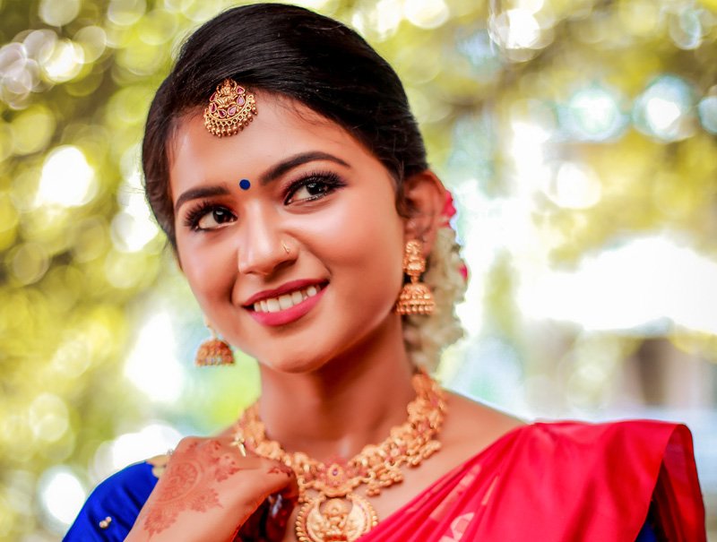 Smiling bride in a red saree with gold jewellery, trees behind her