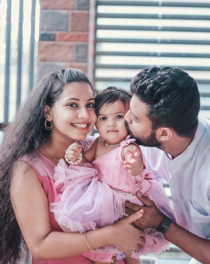 Couple holding their baby daughter in a pink dress, the father kissing her cheek