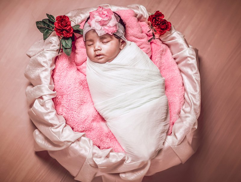 Newborn baby asleep, wrapped in white, in a pink basket decorated with roses