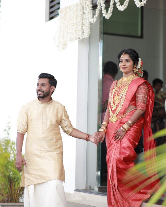 Bride in a red saree and gold jewellery holding hands with the groom outside a house decorated with jasmine